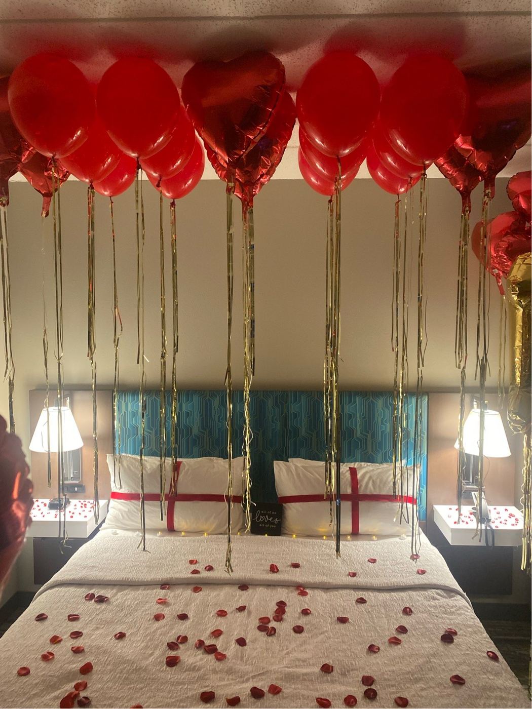 Hotel room styled with a red heart balloon canopy and rose petals across the bed